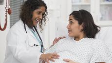 Healthcare worker standing beside a pregnant person lying in bed while both are touching the pregnant person's stomach