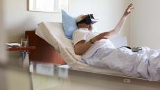 Person in a clinical setting lying in bed with their hand raised while wearing a virtual reality headset