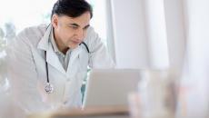 Healthcare provider wearing a lab coat with a stethoscope around their neck while looking at a computer